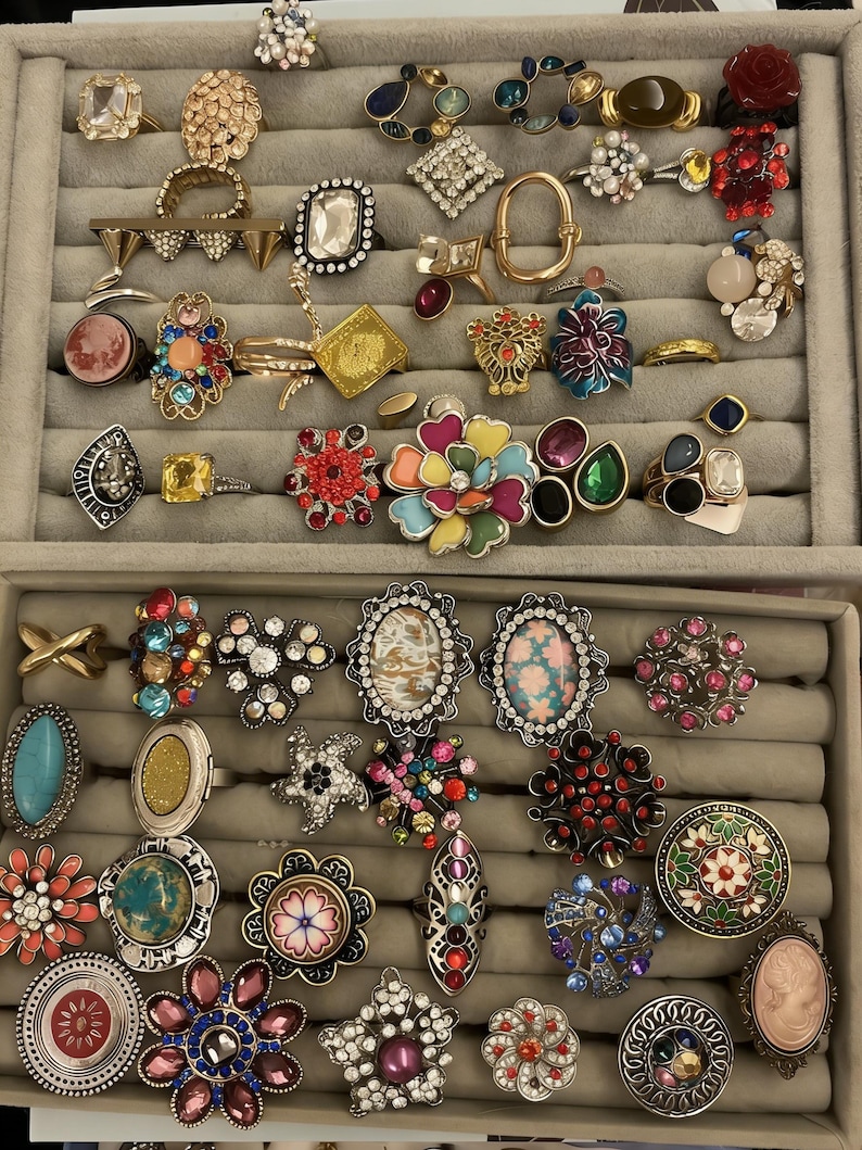 May include: A selection of rings in a grey jewellery tray. The rings have diverse designs, including floral, geometric, and gemstone embellishments. Colours range from vibrant reds and blues to neutral tones, showcasing a variety of styles and materials.