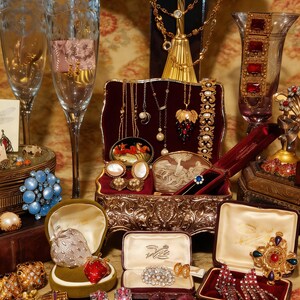 May include: A collection of antique jewellery, including necklaces, brooches, earrings, and rings, presented in open boxes and on a stand. The jewellery features gold, silver, and colourful gemstones. Champagne flutes and decorative vases are also visible.