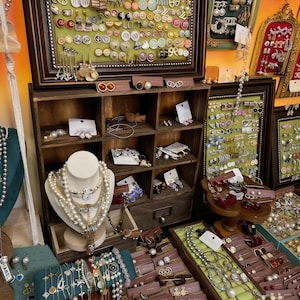 May include: A shop display showcasing a variety of jewellery pieces. Necklaces, earrings, and bracelets are arranged on wooden shelves and display cases. The jewellery features pearls, gold, and silver, in diverse styles and colours.