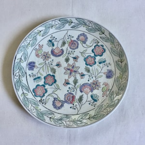 May include: A white ceramic plate with a floral design. The plate features a border of green leaves and a center filled with pink, purple, and blue flowers.