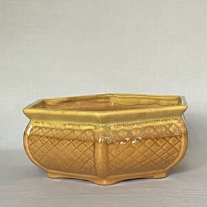 May include: A rectangular ceramic planter with a glossy golden-yellow glaze. The exterior features a raised diamond-patterned texture on the sides, a smooth rim, and four small feet at the base. The finish shows subtle variations in color tone.