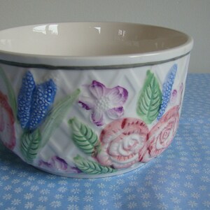 May include: White ceramic bowl with embossed floral design. The bowl features pink, purple, and blue flowers with green leaves against a white background. The rim is gray.