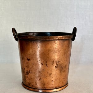 May include: A cylindrical copper bucket featuring a weathered, metallic finish with visible surface marks and patina. The vessel includes two small, dark metal ring handles attached to the upper rim on opposite sides, creating a functional design.