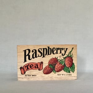 May include: Wooden box of raspberry tea with the text "Raspberry Tea" in black script. The box features a graphic of red raspberries with green leaves. The box contains 25 tea bags and weighs 1.5 oz.