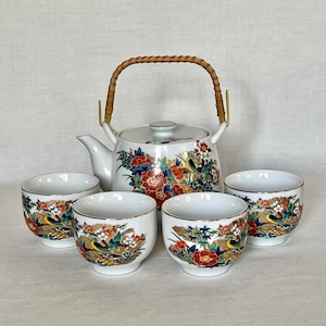 May include: A white porcelain tea set consisting of a teapot with a woven bamboo handle and four matching cups. The set features a colorful pattern of red, blue, and orange flowers, green foliage, and small yellow birds on each piece.