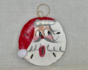 May include: A hand-painted Christmas ornament shaped like Santa Claus's face. The ornament is made from a white sand dollar with a red hat, white beard, and a gold string for hanging. A small white pom-pom is attached to the hat.