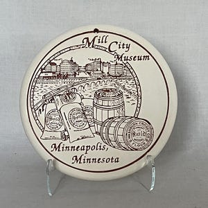 May include: A round, off-white ceramic coaster featuring a brown illustration of the Mill City Museum in Minneapolis, Minnesota. The design includes buildings, a bridge, barrels, and flour sacks. The text "Mill City Museum" and "Minneapolis, Minnesota" are also present.