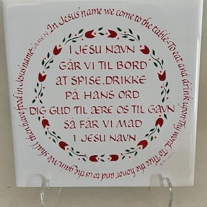 May include: A square white ceramic tile featuring a red floral wreath border and red text in both English and Norwegian. The English text reads: In Jesus' name we come to the table-To eat and drink upon Thy word. To Thee the honor and to us the gain.