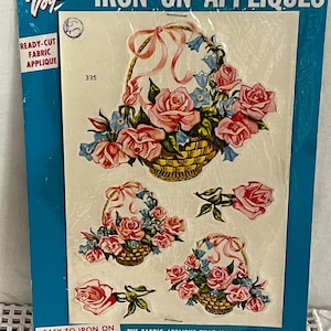 May include: Vintage Vogart iron-on fabric appliqués featuring floral designs. The package includes ready-cut appliqués of pink roses and blue flowers in woven baskets, with additional loose rose and leaf accents. The package is blue and white.