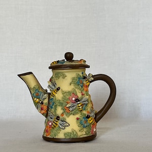 May include: A cream-colored ceramic teapot featuring a raised, three-dimensional relief design of yellow and black bumblebees, green leaves, and small blue and orange flowers. The teapot has a dark brown handle, base, and lid knob.