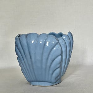 May include: A light blue ceramic planter shaped like a seashell with a glossy finish. The vessel features vertical ridges and a scalloped top edge, creating a textured, shell-like appearance. The base is circular and the interior is hollow.