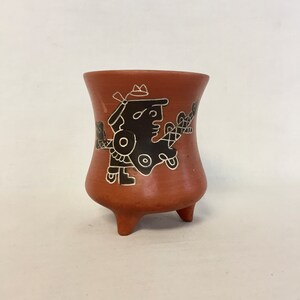 May include: A terracotta pottery cup with three legs and a black and white design of a stylized figure. The figure is wearing a crown and has a large eye.