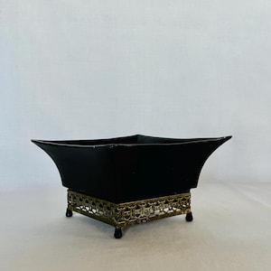 May include: A black, square-shaped decorative bowl with flared sides. The bowl sits on a gold-colored, ornate metal base with small feet. The bowl's design is simple, yet elegant, suitable for various decorative purposes.