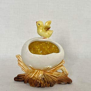 May include: A ceramic Easter decoration featuring a yellow chick perched atop a cracked white egg filled with yellow filler. The egg sits on a brown and tan straw-like base. The chick has a red beak and open wings.