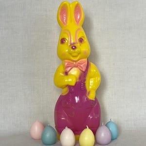 May include: A large, yellow Easter bunny figurine wearing pink overalls and a pink bow tie. Seven pastel-colored egg-shaped candles are arranged in front of the bunny. The bunny is a vintage decoration.