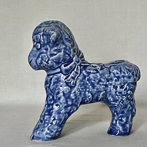 May include: A blue ceramic figurine of a lamb with a textured, curly wool surface and a smooth bow tied around its neck. The figurine features a hollow opening on its back, indicating its function as a small planter or decorative vessel.