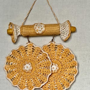 May include: A handmade, wall-mounted kitchen organizer featuring a crocheted design in shades of yellow and white. The organizer includes a rod with two matching doilies hanging below. The piece is suspended by a woven cord.