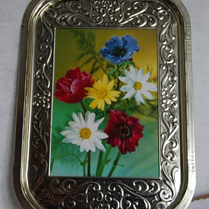 May include: A rectangular metal serving tray with an ornate silver-colored frame. The tray's center features a floral arrangement of red, yellow, white, and blue flowers against a green and yellow background. The tray is approximately 16 inches long and 10 inches wide.
