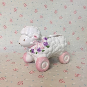 May include: White ceramic lamb planter with pink wheels and purple flowers. The lamb has a pink ribbon around its neck.
