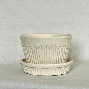 May include: Off-white ceramic planter with a matching saucer. The planter has vertical ribbed detailing and a decorative zigzag pattern near the rim. The saucer also has a ribbed design. The set is suitable for small plants and adds a touch of vintage charm.