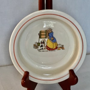 May include: A white ceramic dish with a red rim features a colorful illustration of Old Mother Hubbard and her dog. The illustration shows Old Mother Hubbard standing in a doorway with her dog by her side. The dish is on a wooden stand.