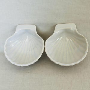 May include: Two white, shell-shaped dishes with a pearlescent sheen. Each dish has a scalloped edge and a small, rectangular handle. The dishes are arranged side by side, showcasing their identical design and iridescent finish.