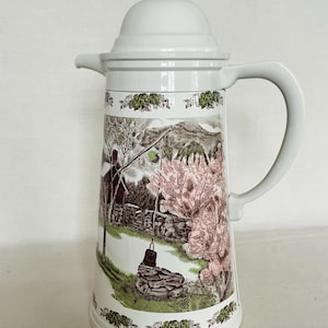 May include: White ceramic thermal carafe with a floral pattern depicting a country scene with a stone wall, a well, and trees. The carafe has a lid with a knob on top.