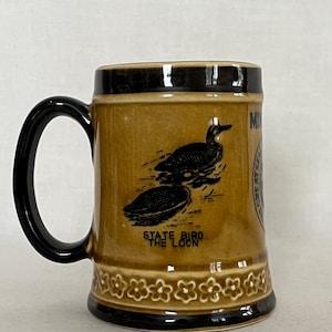 May include: A brown ceramic mug with a black handle and rim. The mug features a black illustration of two loons and the text "STATE BIRD THE LOON". The mug has a floral design around the base and the words "MINNESOTA GREAT SEAL".