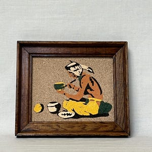 May include: A framed sand art piece on a cork background depicting a person with a white headband holding a green bowl. The figure wears yellow pants and sits near three small bowls. The frame is made of dark brown wood with a visible grain texture.