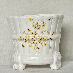 May include: White ceramic planter with a fluted design and three small feet. The planter is decorated with yellow and brown floral accents. A decorative piece for indoor plants.
