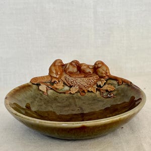 May include: A ceramic dish with a raised relief of birds in a nest. The dish has a shallow, oval shape with a mottled green and brown glaze. The birds and nest are a warm brown color, adding a naturalistic touch to the decorative piece.