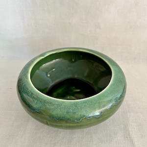 May include: A round ceramic bowl with a glossy finish in shades of deep forest green and lighter sage green. The bowl features a wide, thick rim and a smooth, curved exterior that transitions into a dark, reflective interior basin.