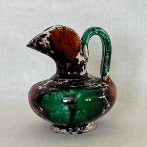 May include: A small, decorative ceramic pitcher with a rounded base and a curved handle. The pitcher features a spout and is adorned with a mix of colors, including green, brown, and black. The handle is a vibrant green, and the overall design is unique.