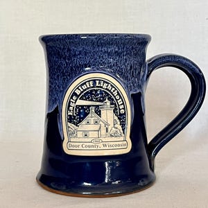 May include: A dark blue ceramic mug with a speckled glaze finish. The front features a cream-colored emblem depicting the Eagle Bluff Lighthouse in Door County, Wisconsin, with the text "Eagle Bluff Lighthouse" and "1868".