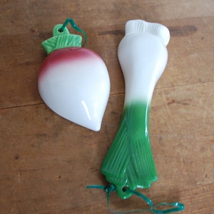 May include: Two ceramic kitchen utensil holders shaped like a turnip and a leek. The turnip is red and white with green leaves. The leek is white and green with a green stem.