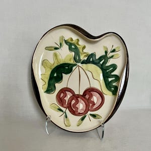 May include: A ceramic, heart-shaped plate with a cream-colored interior and a dark brown rim. The plate features a hand-painted design of three red cherries with green leaves and stems. The plate is displayed on a clear stand.