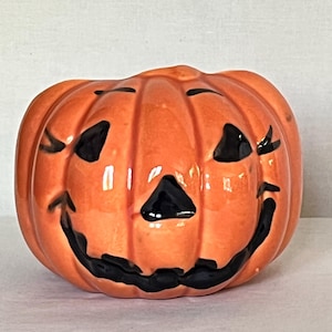 May include: A glossy orange ceramic jack-o'-lantern with a smooth, rounded pumpkin shape. It features a hand-painted black face with triangular eyes, a small triangular nose, and a wide, curved mouth with jagged edges, creating a classic look.