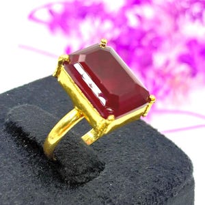 Könnte beinhalten: Ein goldener Ring mit einem großen, rechteckigen, tiefroten Edelstein. Der Ring befindet sich auf einem schwarzen Präsentationsständer, mit einem verschwommenen Hintergrund aus lila Blumen. Der Ring hat ein goldfarbenes Band und eine Fassung.