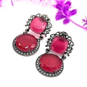 Könnte beinhalten: Ein Paar Ohrhänger mit dunklem Metallrahmen. Jeder Ohrring hat oben einen quadratischen rosa Edelstein und unten einen runden rosa Edelstein. Die Edelsteine sind von kleinen, klaren Kristallen umgeben.