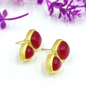 Könnte beinhalten: Ein Paar goldfarbene Ohrstecker, jeweils mit zwei ovalen, tiefroten Edelsteinen. Die Ohrringe haben ein schlichtes, elegantes Design, wobei die Edelsteine in einer goldfarbenen Lünette gefasst sind. Der Hintergrund ist weiß mit lila floralen Akzenten.