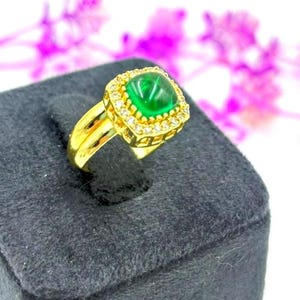 Könnte beinhalten: Ein goldener Ring mit einem quadratischen, smaragdgrünen Edelstein in der Mitte. Der Edelstein ist von einem Kranz aus kleinen, klaren Steinen umgeben. Der Ring wird auf einer schwarzen Samtoberfläche mit verschwommenem floralen Hintergrund präsentiert.