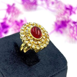 Könnte beinhalten: Ein goldener Ring mit einem großen, ovalen, roten Edelstein in der Mitte, umgeben von kleineren, klaren Edelsteinen. Der Ring hat ein detailliertes, verziertes Design. Er wird auf einer schwarzen Samtoberfläche mit verschwommenem floralen Hintergrund präsentiert.