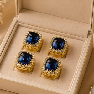 Könnte beinhalten: Ein Set aus vier quadratischen Manschettenknöpfen mit einem tiefblauen Cabochon-Zentrum, umgeben von einer Umrandung aus kleinen klaren Steinen in einem goldfarbenen Metallrahmen, präsentiert in einer beigen rechteckigen Schachtel mit Klappdeckel.