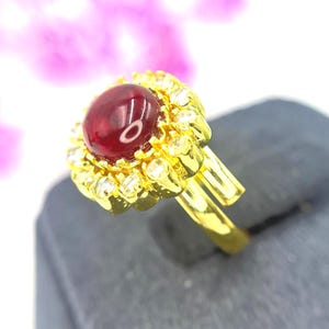 Könnte beinhalten: Ein goldener Ring mit einem großen, ovalen, tiefroten Edelstein in der Mitte, umgeben von kleineren, klaren Edelsteinen. Der Ring und die Fassung sind in einem hellen, polierten Goldton gehalten. Der Ring wird vor einem weichen, verschwommenen Hintergrund gezeigt.