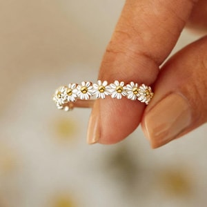May include: A silver-toned ring featuring a continuous band of small daisy flowers. Each flower has white petals and a small, raised gold-toned center. The ring is designed as a circular band with a repeating floral pattern around the entire circumference.