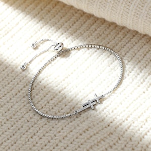 May include: A silver bracelet with a cross pendant. The bracelet features a delicate chain and a sliding clasp for adjustable sizing. The cross is embellished with small, sparkling stones, adding a touch of elegance to the jewelry.