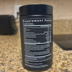 May include: A black supplement bottle with white text detailing the supplement facts. The label lists ingredients such as Vitamin A, Vitamin C, Vitamin D, and other vitamins and minerals. The bottle is on a light-colored countertop.