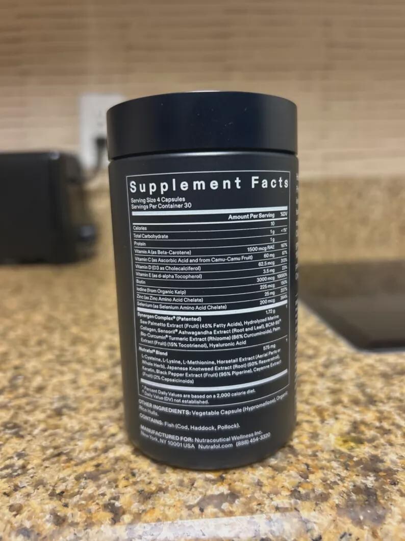 May include: A black supplement bottle with white text detailing the supplement facts. The label lists ingredients such as Vitamin A, Vitamin C, Vitamin D, and other vitamins and minerals. The bottle is on a light-colored countertop.