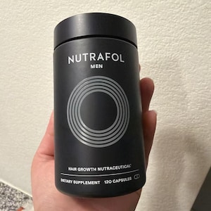 May include: A black cylindrical bottle of Nutrafol Men hair growth nutraceutical dietary supplement. The bottle has a black cap and features the brand name and product information in white text. The bottle contains 120 capsules.