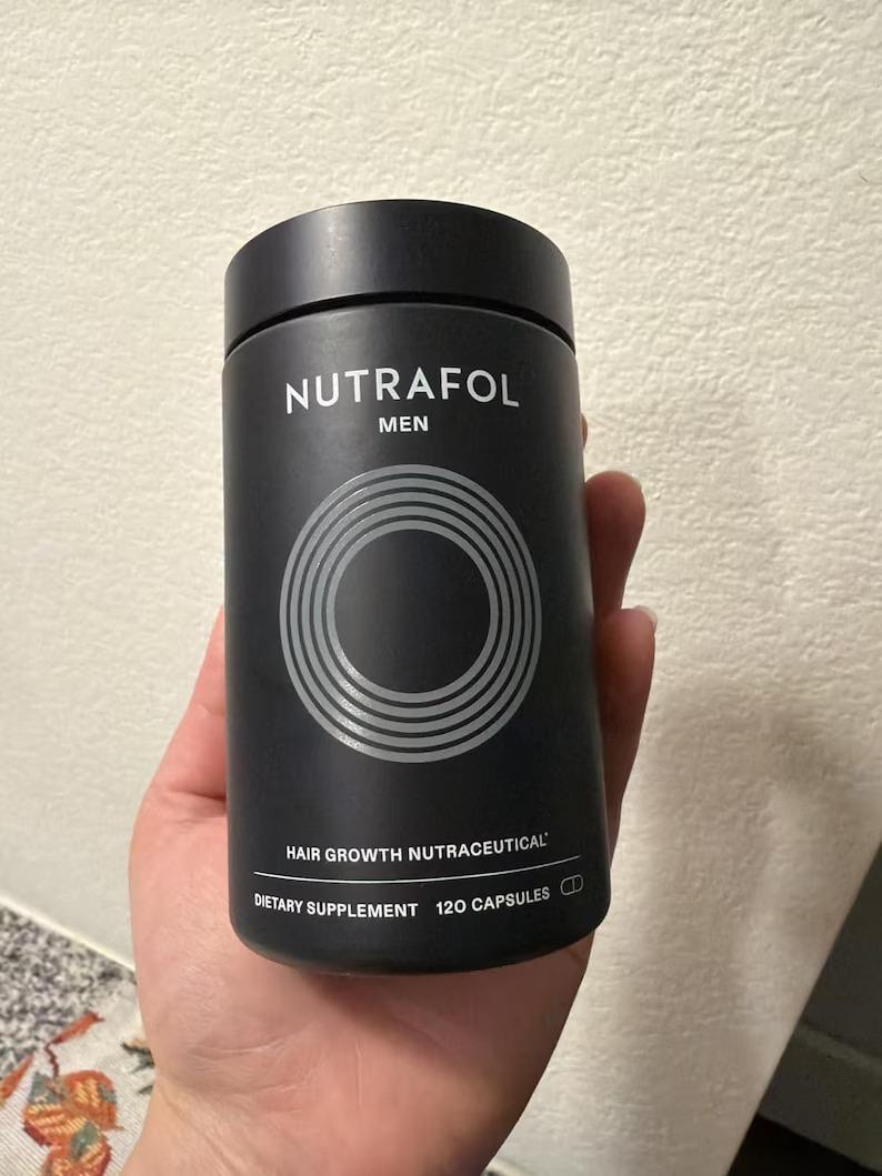 May include: A black cylindrical bottle of Nutrafol Men hair growth nutraceutical dietary supplement. The bottle has a black cap and features the brand name and product information in white text. The bottle contains 120 capsules.
